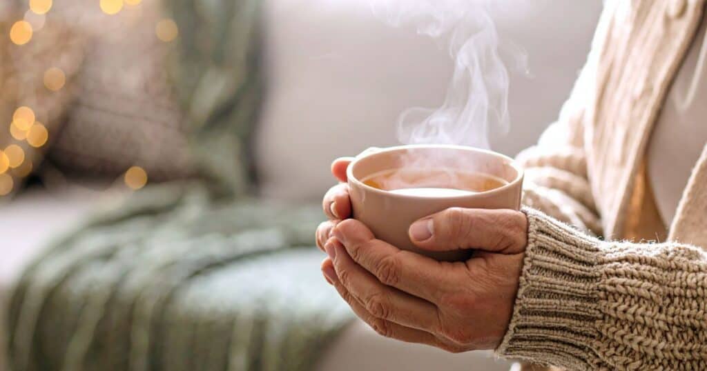 Woman sitting beside a window holding a warm mug of tea in her hands for comfort